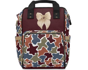 May include: A backpack featuring a deep burgundy upper section with a beige bow graphic. The lower front pocket and side panels display a multi-colored abstract camouflage pattern in shades of burgundy, blue, olive green, and white with black trim.