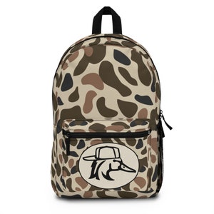 May include: A camouflage-patterned backpack in shades of tan, brown, and black. The backpack features a front pocket with a zipper, a black top handle, and a side mesh pocket. A circular patch with a duck wearing a hat is on the front.