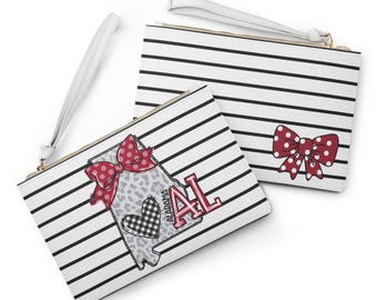 May include: Two white wristlet pouches featuring horizontal black stripes. One pouch displays a red polka-dot bow, while the other features a gray leopard-print Alabama state outline, a black and white checkered heart, and the red text ALABAMA AL.
