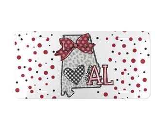 May include: A rectangular license plate featuring a white background with red and black polka dots. The center displays a gray leopard print outline of the state of Alabama, a red bow, a black and white buffalo plaid heart, and the text ALABAMA AL.