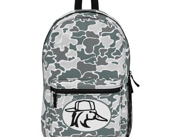 May include: A backpack with a camouflage pattern in shades of gray and white. The backpack features a black top handle, a front zippered pocket, and a side mesh pocket. A white oval with a black duck logo is centered on the front.