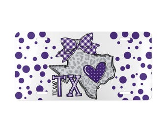 May include: A rectangular license plate featuring a white background with purple polka dots. The center displays a gray leopard print outline of the state of Texas, a purple gingham bow, a purple heart with white dots, and the text TEXAS TX in purple.