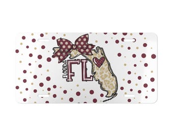 May include: A rectangular license plate with a white background featuring maroon and gold polka dots. The center displays a leopard print outline of the state of Florida with the text FLORIDA FL, a maroon bow, and a small maroon heart with white dots.