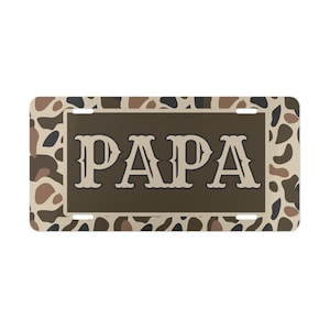 May include: Rectangular license plate with a camouflage pattern in shades of brown, tan, and black. The word "PAPA" is displayed in large, stylized, off-white letters on a brown background.