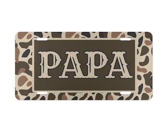 May include: Rectangular license plate with a camouflage pattern in shades of brown, tan, and black. The word "PAPA" is displayed in large, stylized, off-white letters on a brown background.