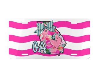 May include: A rectangular license plate featuring horizontal hot pink and white wavy stripes. The center displays a floral-patterned outline of the state of Georgia with a light blue striped bow, a pink polka-dot heart, and the text GA and GEORGIA.