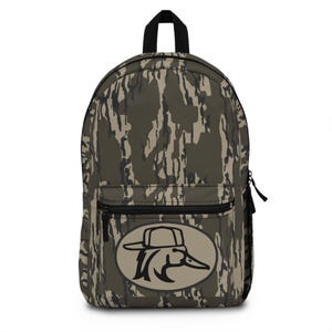May include: A camouflage-patterned backpack with a black top handle, zipper, and side pocket. The front features a circular emblem with a black duck wearing a hat. The backpack is designed for carrying items.