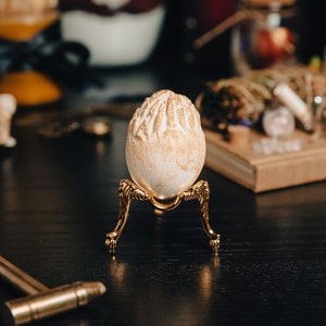 K&ouml;nnte beinhalten: Ein wei&szlig;es Ei mit strukturierter Oberfl&auml;che steht auf einem goldenen Metallst&auml;nder mit drei Beinen. Der St&auml;nder steht auf einer schwarzen Oberfl&auml;che.