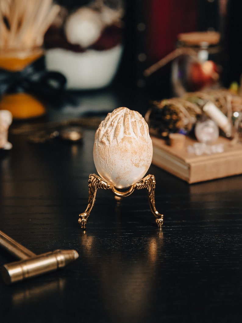 K&ouml;nnte beinhalten: Ein wei&szlig;es Ei mit strukturierter Oberfl&auml;che steht auf einem goldenen Metallst&auml;nder mit drei Beinen. Der St&auml;nder steht auf einer schwarzen Oberfl&auml;che.