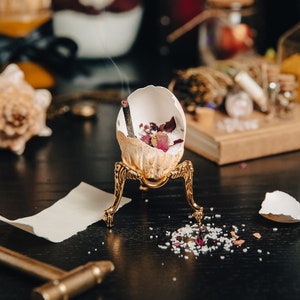 K&ouml;nnte beinhalten: Eine wei&szlig;e Eierschale mit getrockneten Rosenbl&uuml;ten und einem glimmenden R&auml;ucherst&auml;bchen steht auf einem goldenen Metallst&auml;nder. Der St&auml;nder hat drei Beine und steht auf einer schwarzen Oberfl&auml;che. Kleine wei&szlig;e und rosa Kristalle sind um den St&auml;nder herum verstreut.