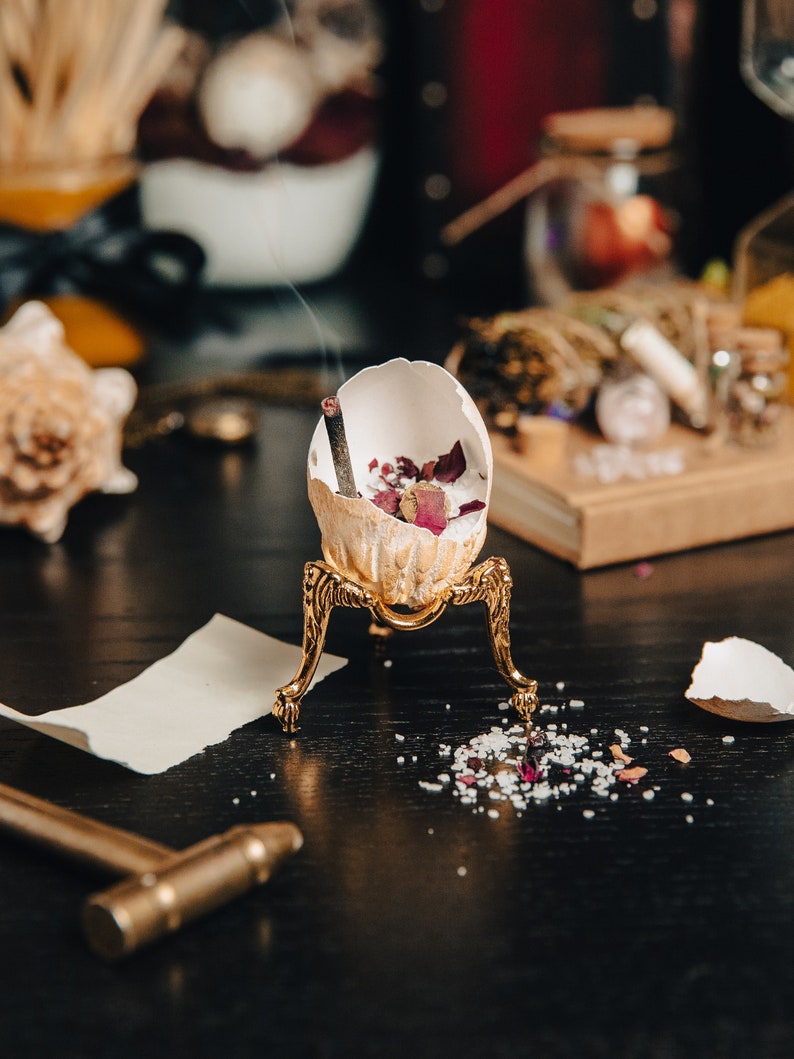 K&ouml;nnte beinhalten: Eine wei&szlig;e Eierschale mit getrockneten Rosenbl&uuml;ten und einem glimmenden R&auml;ucherst&auml;bchen steht auf einem goldenen Metallst&auml;nder. Der St&auml;nder hat drei Beine und steht auf einer schwarzen Oberfl&auml;che. Kleine wei&szlig;e und rosa Kristalle sind um den St&auml;nder herum verstreut.