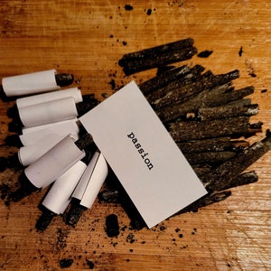 K&ouml;nnte beinhalten: Eine wei&szlig;e Karte mit dem Wort "Passion" in schwarzer Schrift, die auf einer Holzoberfl&auml;che liegt, die mit schwarzen Holzkohle-St&auml;bchen bedeckt ist.