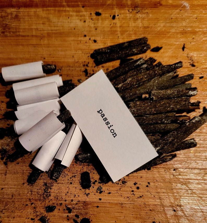 K&ouml;nnte beinhalten: Eine wei&szlig;e Karte mit dem Wort "Passion" in schwarzer Schrift, die auf einer Holzoberfl&auml;che liegt, die mit schwarzen Holzkohle-St&auml;bchen bedeckt ist.