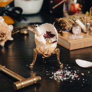 Peut inclure: Un support en métal doré à trois pieds soutient une coquille d'œuf blanche contenant un morceau de papier roulé, des pétales de fleurs séchées et une perle dorée. Le support est posé sur une surface noire avec un petit marteau et des cristaux blancs et roses éparpillés.