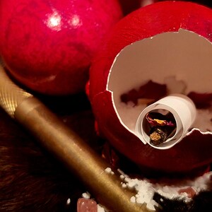 K&ouml;nnte beinhalten: Ein rotes, rissiges Ei mit einem wei&szlig;en Papierrohr im Inneren. Das Rohr enth&auml;lt getrocknete Blumen und ein kleines St&uuml;ck Holz. Das Ei liegt auf einem goldenen Tablett mit wei&szlig;en Streuseln und rosa Bl&uuml;tenbl&auml;ttern.
