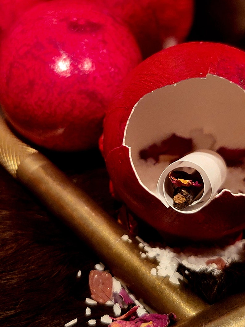 K&ouml;nnte beinhalten: Ein rotes, rissiges Ei mit einem wei&szlig;en Papierrohr im Inneren. Das Rohr enth&auml;lt getrocknete Blumen und ein kleines St&uuml;ck Holz. Das Ei liegt auf einem goldenen Tablett mit wei&szlig;en Streuseln und rosa Bl&uuml;tenbl&auml;ttern.