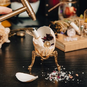 K&ouml;nnte beinhalten: Ein goldfarbener, eif&ouml;rmiger Beh&auml;lter mit einer kleinen goldenen Kugel im Inneren, der auf einem dreibeinigen St&auml;nder steht. Das Ei ist aufgebrochen und zeigt ein aufgerolltes St&uuml;ck Papier und ein paar rote Rosenbl&auml;tter. Ein kleiner Hammer schl&auml;gt auf das Ei.