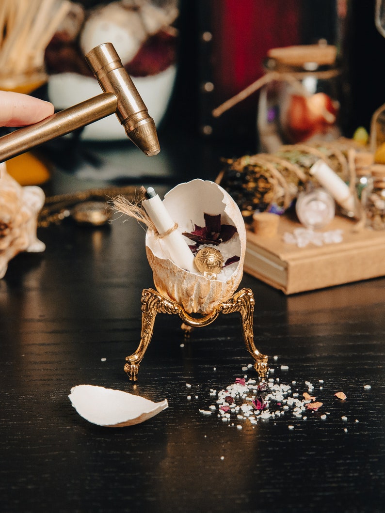 K&ouml;nnte beinhalten: Ein goldfarbener, eif&ouml;rmiger Beh&auml;lter mit einer kleinen goldenen Kugel im Inneren, der auf einem dreibeinigen St&auml;nder steht. Das Ei ist aufgebrochen und zeigt ein aufgerolltes St&uuml;ck Papier und ein paar rote Rosenbl&auml;tter. Ein kleiner Hammer schl&auml;gt auf das Ei.