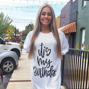 May include: A white sequined dress with the text "It's my Birthday" in black script. The dress is paired with silver glitter cowboy boots. The image is taken outdoors on a sidewalk.