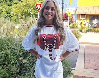 May include: A woman in a white sequined dress featuring a red elephant design and red and white striped trim. She wears silver glitter cowboy boots. The background shows greenery and buildings.