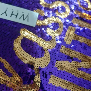 May include: A fabric surface covered in purple and gold sequins, featuring the text "WHY DRESS" printed on a white rectangular tag. The sequins are arranged to form letters in gold against a purple background, creating a shimmering texture.
