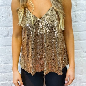 May include: A gold sequin camisole top with a V-neck. The top is sleeveless and shimmers with a multitude of small, reflective sequins. The model is wearing black pants. The background is a white brick wall.