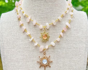 Puede incluir: Un collar de varias capas con piedras de cuarzo rosa y dos colgantes de sol dorados. El collar está en un maniquí de tela blanca.