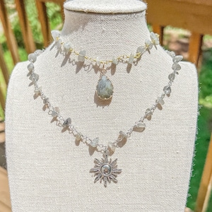 Puede incluir: Dos collares superpuestos con cadenas de plata y oro. El collar superior tiene un colgante en forma de lágrima con una piedra verde claro. El collar inferior tiene un colgante de sol plateado.