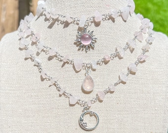 Puede incluir: Tres collares superpuestos con cuentas de piedra rosa y cadena plateada. El collar superior tiene un dije de sol plateado, el collar del medio tiene un dije de lágrima plateada y el collar inferior tiene un dije de luna creciente plateada.