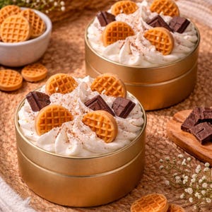 Puede incluir: Dos latas doradas llenas de crema batida, mini gofres y trozos de chocolate. Los postres están espolvoreados con un polvo marrón y se encuentran sobre una superficie tejida. Un pequeño cuenco de mini gofres está en el fondo.
