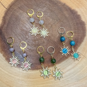 Puede incluir: Surtido de pendientes de tono dorado con colgantes de sol y piedras preciosas. Los pendientes presentan una variedad de colores, incluyendo turquesa, verde, rosa y blanco.