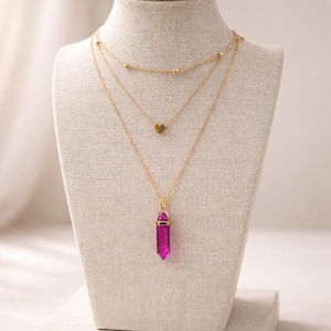 Puede incluir: Conjunto de tres collares expuestos en un maniquí. La cadena superior tiene pequeñas cuentas doradas, la del medio tiene un colgante de corazón dorado y la inferior presenta una cadena dorada con un colgante de cristal rosa.