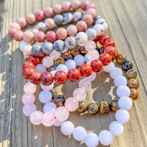 Puede incluir: Varios brazaletes de cuentas en varios colores, incluyendo rosa, rojo, blanco y marrón. Los brazaletes están hechos de piedras redondas y pulidas y están dispuestos sobre una superficie de madera. Los brazaletes son una mezcla de diferentes tipos de piedras.