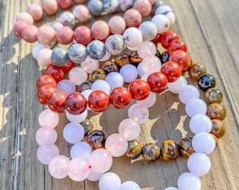May include: Several beaded bracelets in various colors, including pink, red, white, and brown. The bracelets are made of round, polished stones and are arranged on a wooden surface. The bracelets are a mix of different stone types.