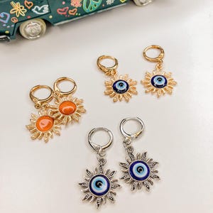 May include: Four pairs of earrings with sun and eye designs. Two pairs have gold-colored hoops and suns in orange and gold. The other two pairs have silver-colored hoops and suns with blue and white eye details.