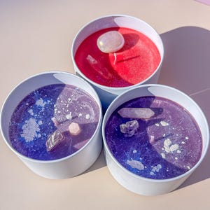 Puede incluir: Tres velas con cristales incrustados en recipientes blancos. Las velas son rojas, moradas y de un tono morado más claro.