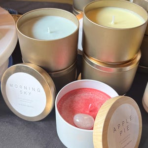 Puede incluir: Se exhiben varias velas perfumadas en latas doradas. Las velas son de varios colores, incluyendo azul claro, amarillo y rojo. Una vela tiene un cristal de cuarzo rosa. Las etiquetas dicen "Morning Sky" y "Apple Pie".