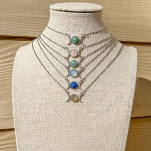 Puede incluir: Un conjunto de siete collares de triple luna plateados con colgantes de piedras preciosas de diferentes colores. Los collares se muestran en un maniquí blanco.