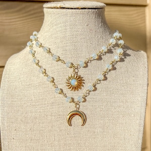 Puede incluir: Un collar dorado de capas con cuentas blancas y dos dijes: un sol con un centro de ópalo y una luna creciente.