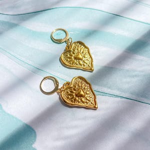 Könnte beinhalten: Ein Paar goldfarbene herzförmige Ohrringe. Jeder Ohrring hat eine strukturierte Oberfläche mit einem Sonnenstrahl-Design in der Mitte. Die Ohrringe haben einen Creolenverschluss und werden auf einer blau-weiß gemusterten Oberfläche präsentiert.