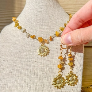 Puede incluir: Un conjunto de collar y pendientes dorados. El collar presenta pequeñas piedras amarillas y blancas y un colgante de girasol. Los pendientes tienen un diseño similar, con un dije de girasol y detalles de piedras. Las joyas se exhiben sobre una tela neutra.