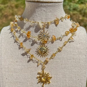 Puede incluir: Un collar de oro con tres capas de pequeñas piedras de color marrón claro y tres colgantes de sol de oro. El collar está en un maniquí blanco.