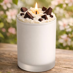 Puede incluir: Una vela de vidrio esmerilado blanco con una mecha encendida. La vela está cubierta con cera blanca y trozos de chocolate negro. La vela está sobre una superficie de madera con un fondo floral borroso.