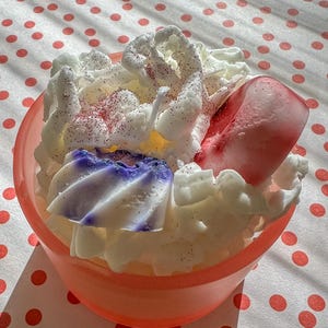 Puede incluir: Una vela decorativa en un cuenco color melocotón, que se asemeja a un postre. La vela presenta cera blanca, similar a crema batida, una pieza de cera roja y blanca y una pieza de cera azul y blanca. Las chispas de purpurina roja añaden detalles.