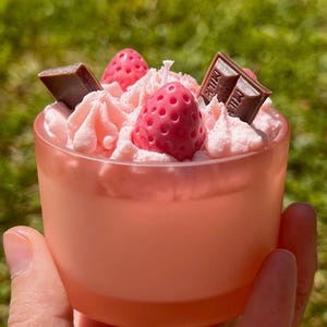 Puede incluir: Una vela rosa en un recipiente de vidrio transparente, decorada para parecerse a un postre de fresa. La vela presenta glaseado rosa, trozos de cera con forma de fresa y detalles de barra de chocolate. Se puede ver una pequeña mecha de vela blanca.