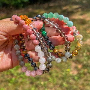 Puede incluir: Una colección de pulseras de cuentas en varios colores, incluyendo rosa, naranja, verde y marrón. Las pulseras están hechas de piedras preciosas pulidas y se sostienen en una mano sobre un fondo verde borroso.