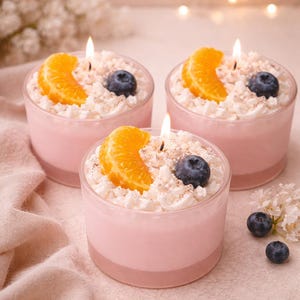 Puede incluir: Tres velas rosas en recipientes de vidrio transparente, cada una coronada con cera blanca, una rodaja de mandarina y un arándano. Las velas están encendidas y la escena está ambientada sobre una superficie rosa claro con flores decorativas.