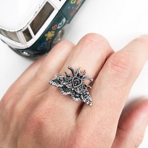 Puede incluir: Un anillo plateado con forma de polilla, con intrincados detalles en las alas y el cuerpo. El anillo se lleva en un dedo, sobre un fondo blanco. Una furgoneta de juguete verde y blanca se encuentra en el fondo.