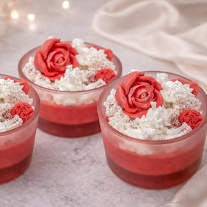 Puede incluir: Tres pequeños vasos transparentes llenos de postres rojos y blancos en capas, cubiertos con decoraciones en forma de rosa roja y crema batida blanca. Los postres están dispuestos sobre una superficie clara.