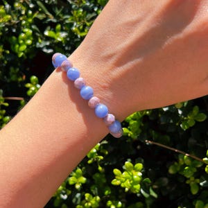 Puede incluir: Un brazalete de cuentas con piedras preciosas azules y rosas alternas. El brazalete se muestra en un brazo, con follaje verde en el fondo. Las piedras preciosas redondas y facetadas crean un accesorio delicado y colorido. Aproximadamente 18-20 cm de largo.